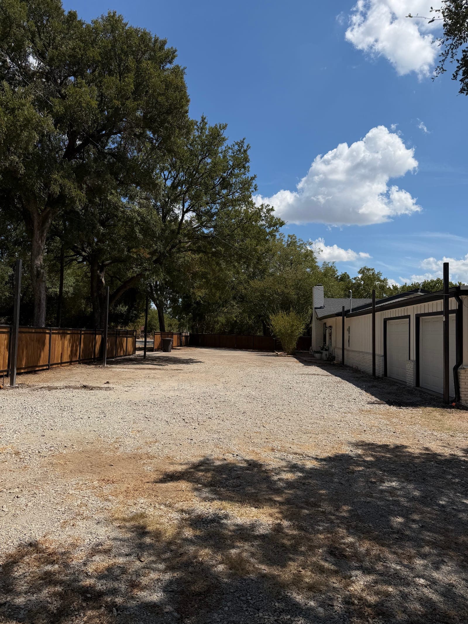 Before: an empty gravel drive between a white house with garage doors and a wood fence under oak trees